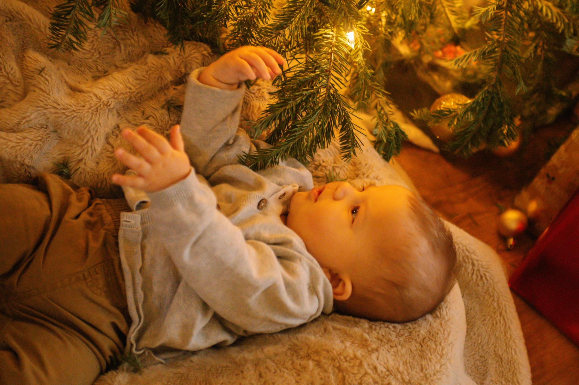Baby lying under a lit Christmas tree, reaching up for a branch in warm lamplight