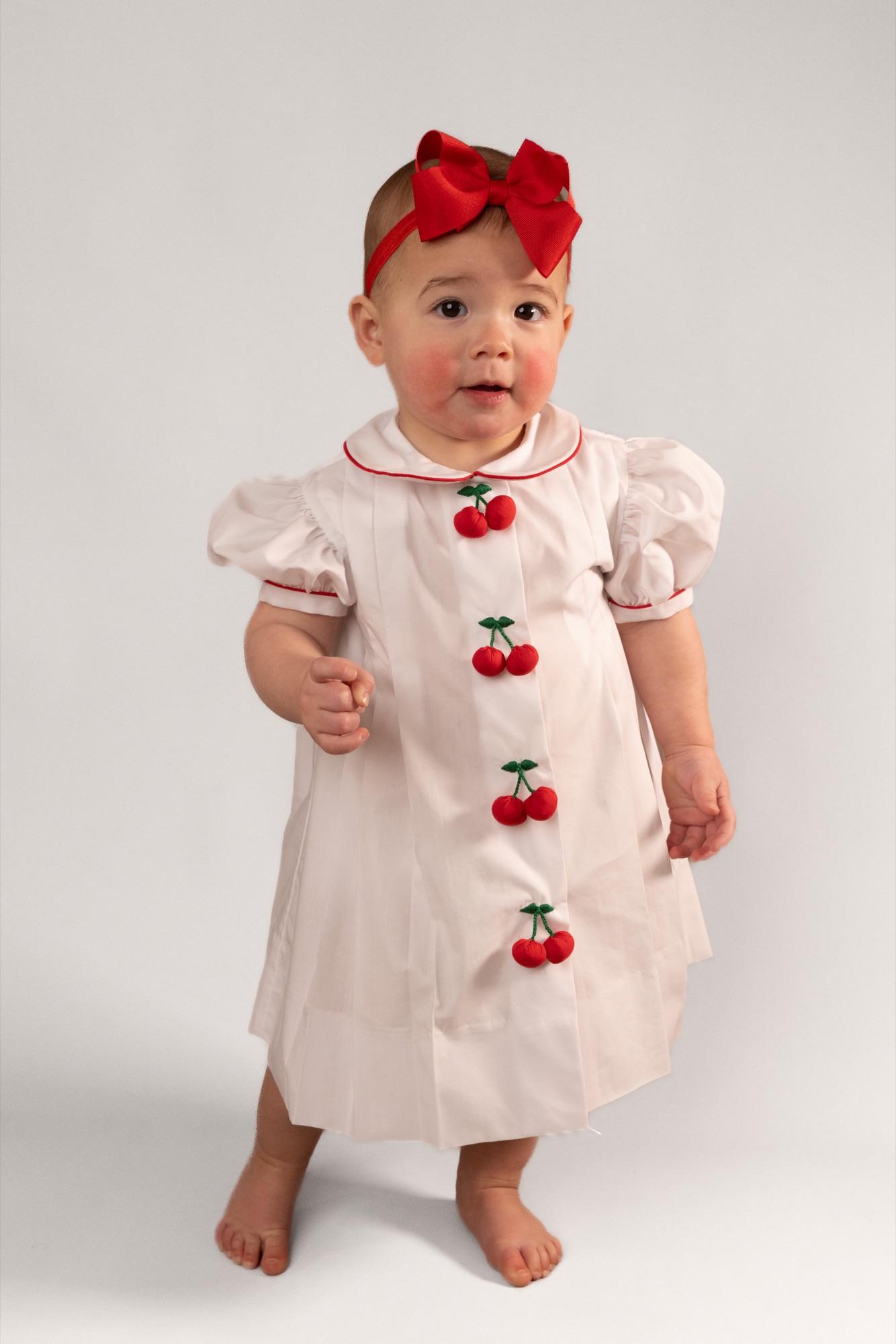 Baby standing in a white cherry-appliqué dress and red bow, full-length on seamless white