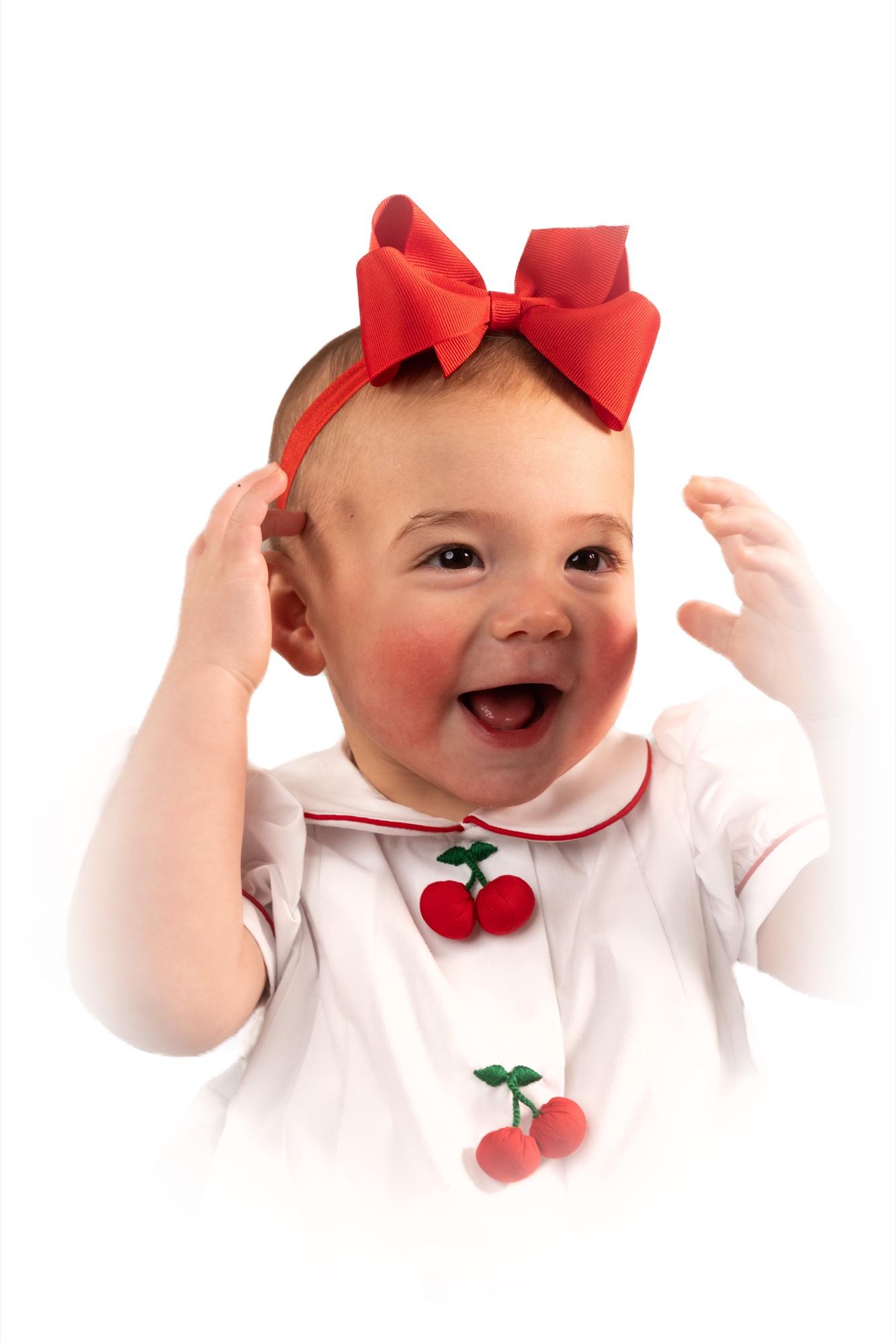 Baby in a red bow throwing both hands up mid-laugh against a white backdrop