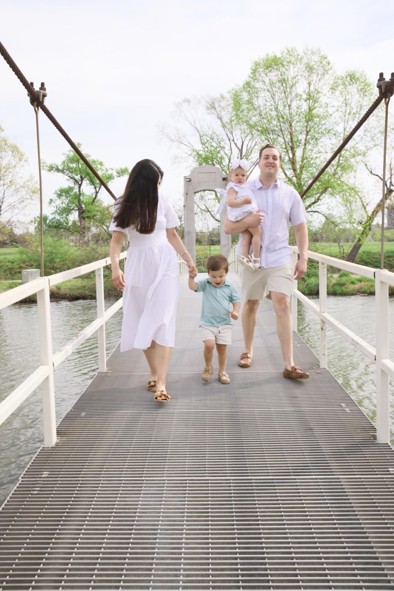 Family of four crossing a park suspension bridge, toddler mid-stride holding hands