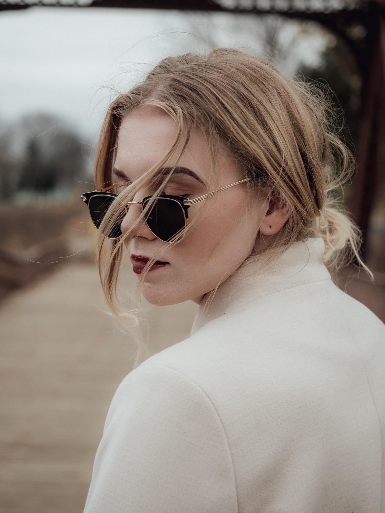 Wind-blown portrait in sunglasses and a white coat on a wooden bridge