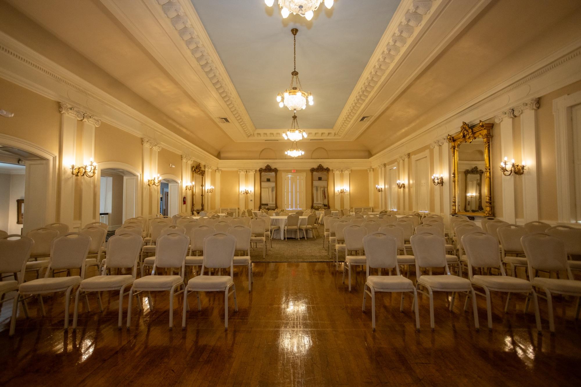 Historic ballroom set for an event — chandeliers, gilded mirrors, and rows of white chairs